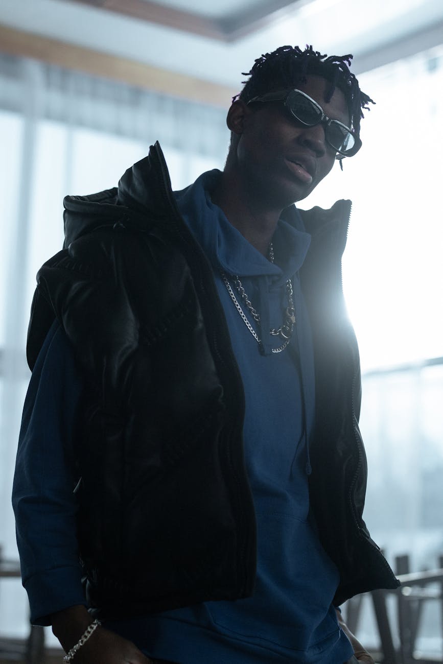 Portrait de mode d'un homme vêtu d'un gilet noir et d'un sweat à capuche bleu posant à l'intérieur avec des lunettes de soleil.